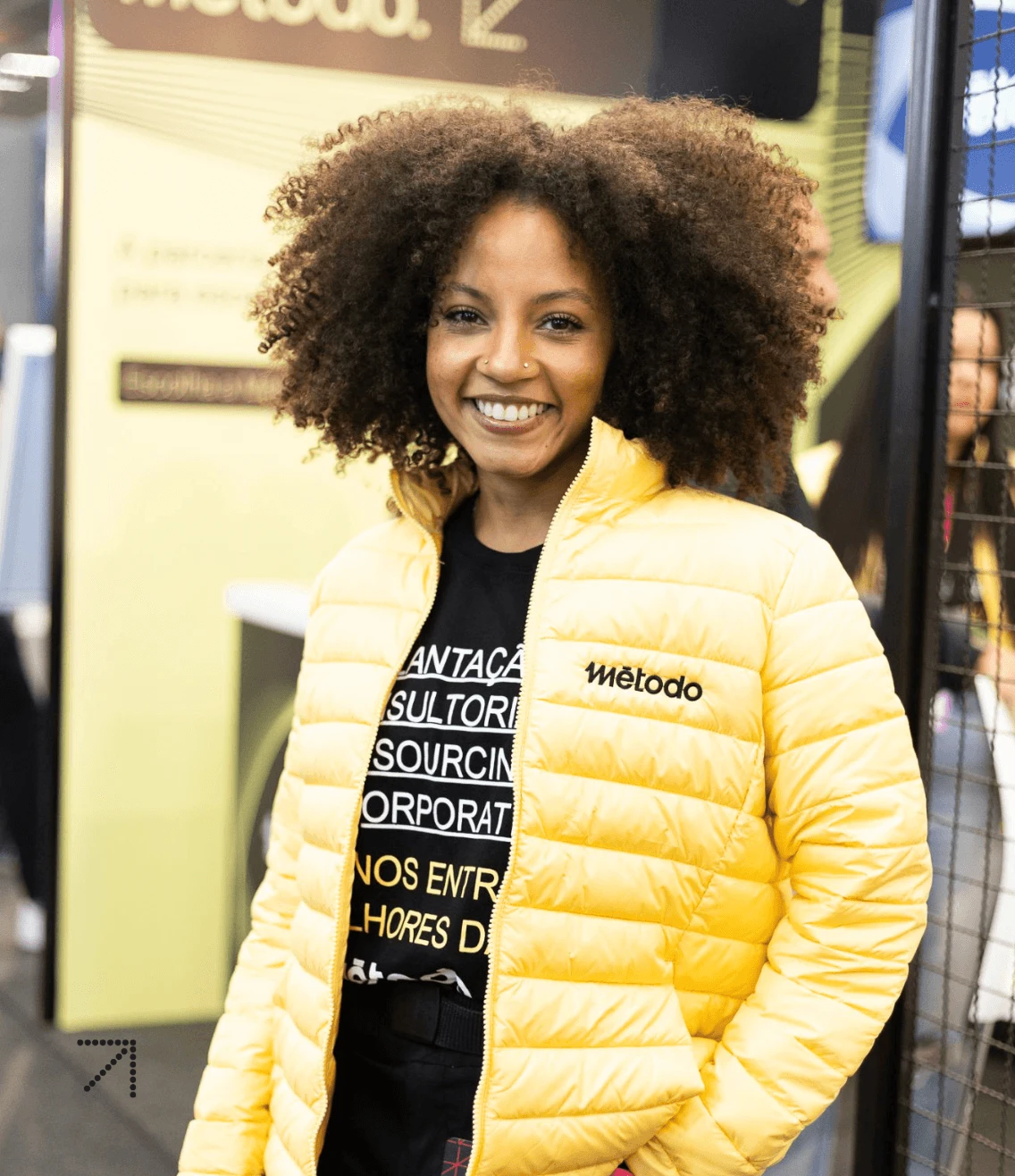 Colaborador Método, vestindo uma jaqueta amarela brilhante e com cabelo cacheado, em pé em um ambiente interno animado.