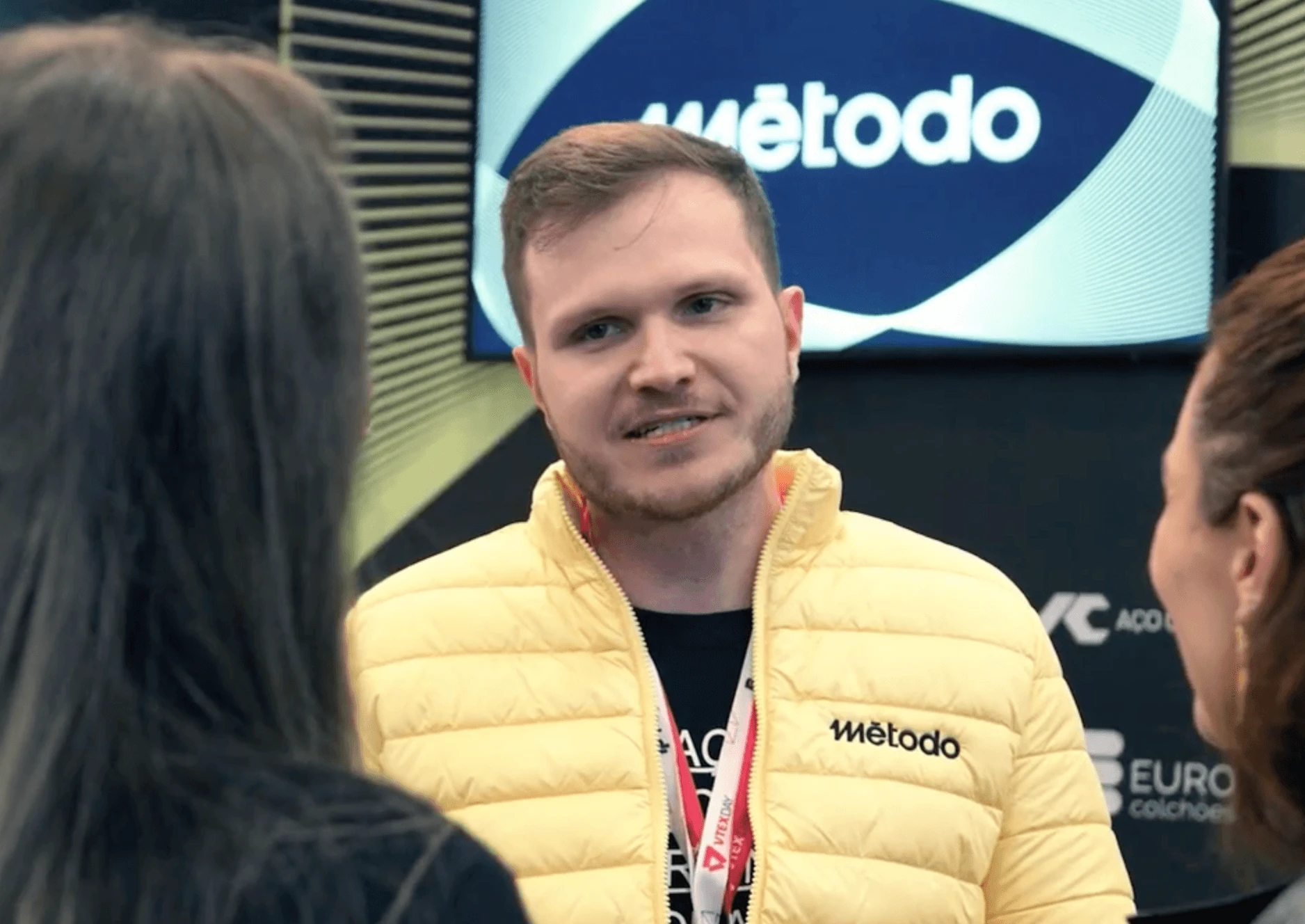 Matheus da Método de jaqueta amarela fala com duas pessoas no VtexDay, com o Stande da fundo.